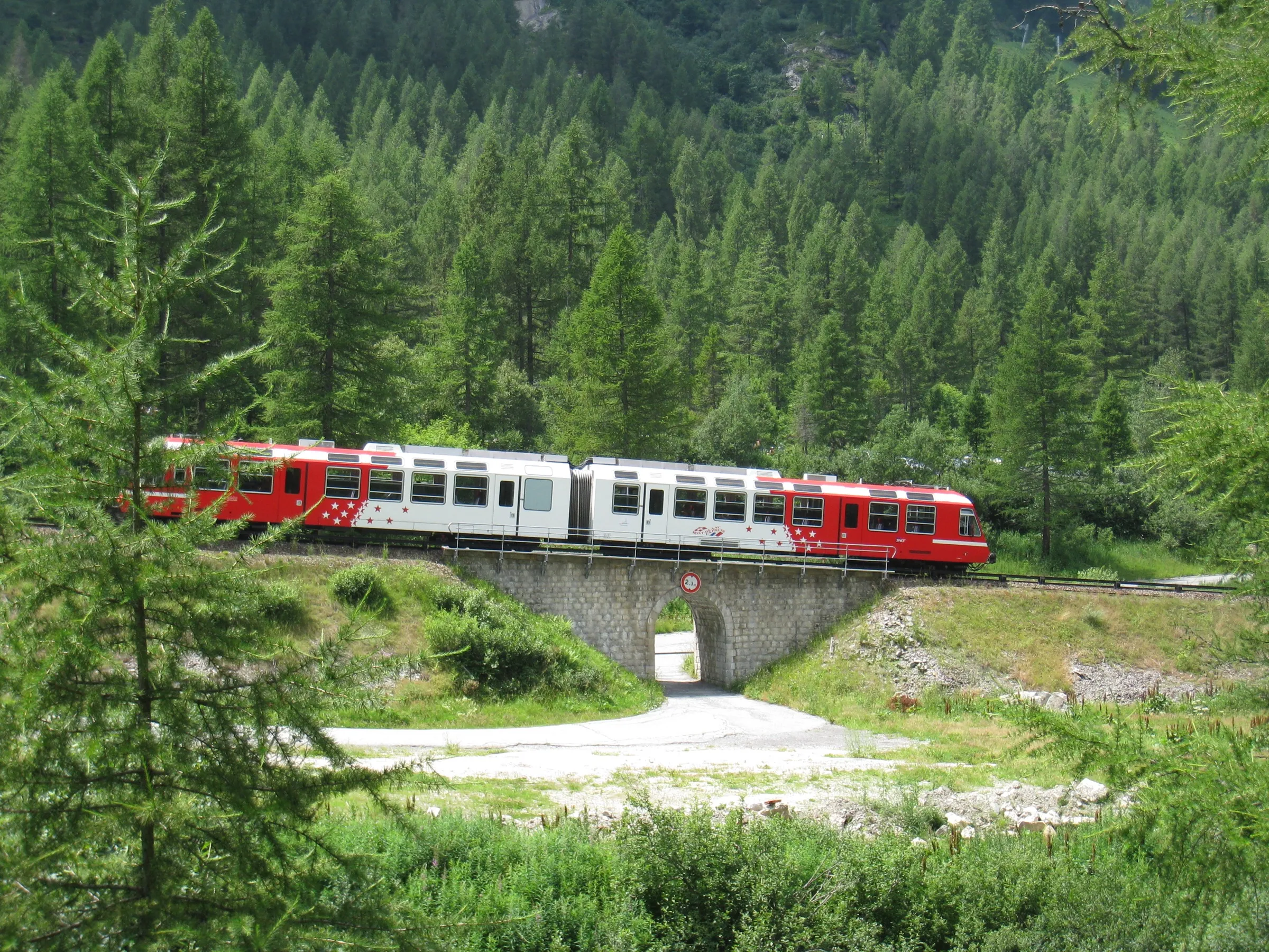 A red and white Mont-Blanc Express train crossing a small stone bridge in a larch forest near Vallorcine.